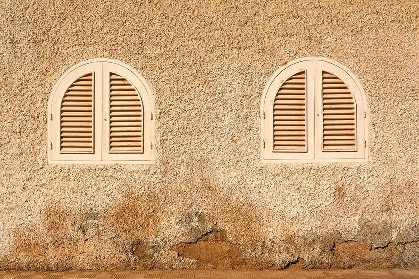 Mur en enduit ocre usé au pied, percé de deux fenêtres à volets cintrés